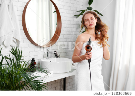 Woman drying her hair with a hair dryer in a modern bathroom with plants, mirror, and white decor Woman drying her hair with a hair dryer in a modern bathroom with plants, mirror, and white decor 128363669