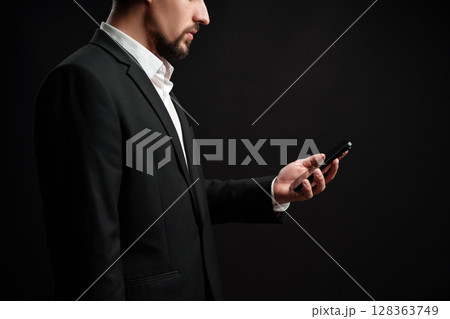 Professional man checking his smartphone while dressed in formal attire against a dark background Professional man checking his smartphone while dressed in formal attire against a dark background 128363749