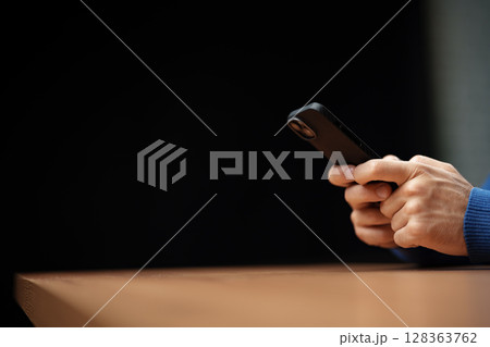 Person using a smartphone while sitting at a wooden table in a dimly lit indoor space during evening hours Person using a smartphone while sitting at a wooden table in a dimly lit indoor space during evening hours 128363762