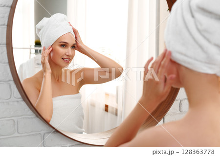 Woman in a towel preparing for the day while admiring herself in a mirror in a bright bathroom at morning time 128363778