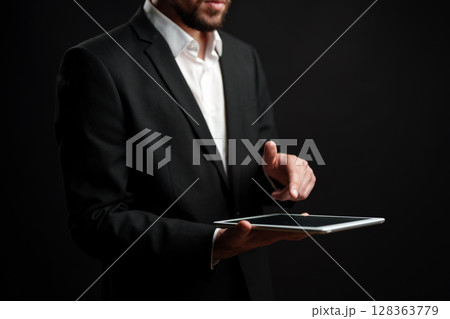 Professional man in suit using tablet while standing against dark background indoors Professional man in suit using tablet while standing against dark background indoors 128363779