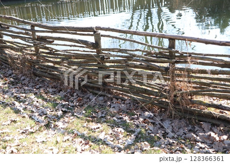 A Rustic Woven Fence standing proudly by the edge of a Tranquil Water body, surrounded by nature A Rustic Woven Fence standing proudly by the edge of a Tranquil Water body, surrounded by nature 128366361