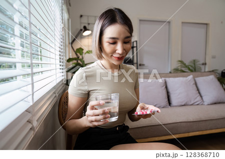Young Woman Taking Medication with Water in a Modern Living Room Setting, Promoting Health and Wellness Young Woman Taking Medication with Water in a Modern Living Room Setting, Promoting Health and Wellness 128368710