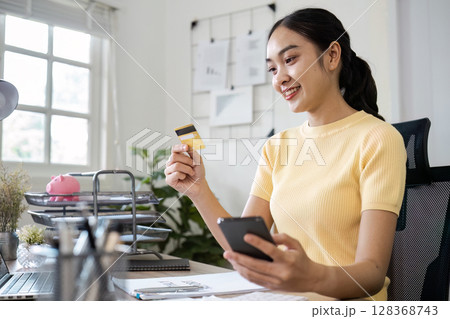 Young Woman Using Credit Card and Mobile Phone for Online Shopping in Modern Home Office Setting 128368743