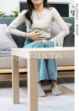 Young Woman Holding Heating Pad on Stomach While Sitting on Couch, Experiencing Menstrual Cramps and Discomfort at Home Young Woman Holding Heating Pad on Stomach While Sitting on Couch, Experiencing Menstrual Cramps and Discomfort at Home 128368754