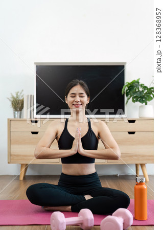 Young Woman Practicing Yoga at Home for Wellness and Relaxation with Fitness Equipment and Plants in the Background Young Woman Practicing Yoga at Home for Wellness and Relaxation with Fitness Equipment and Plants in the Background 128368957