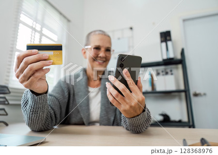 Senior Woman Using Credit Card and Smartphone for Online Shopping in Modern Home Office Setting Senior Woman Using Credit Card and Smartphone for Online Shopping in Modern Home Office Setting 128368976
