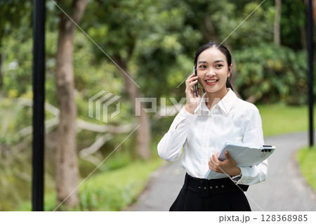 Young Professional Woman Talking on Phone and Holding Documents in Outdoor Park Setting 128368985