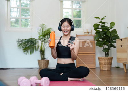 Young Woman Exercising at Home with Dumbbells, Water Bottle, and Smartphone in a Bright Living Room 128369116