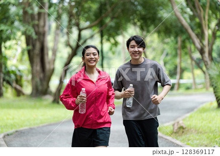 Young Couple Jogging in Park, Smiling and Holding Water Bottles, Healthy Lifestyle Concept 128369117