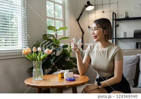Young Woman Taking Medication at Home, Sitting on Sofa with Glass of Water, Surrounded by Healthcare Products and Natural Light 128369340