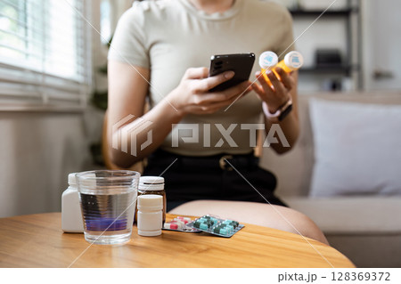 Woman Checking Medication Information on Smartphone While Holding Pill Bottles at Home Woman Checking Medication Information on Smartphone While Holding Pill Bottles at Home 128369372