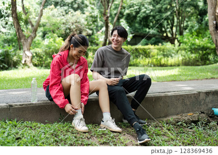 Young Couple Enjoying a Relaxing Jog in the Park, Taking a Break to Laugh and Tie Shoes on a Sunny Day 128369436