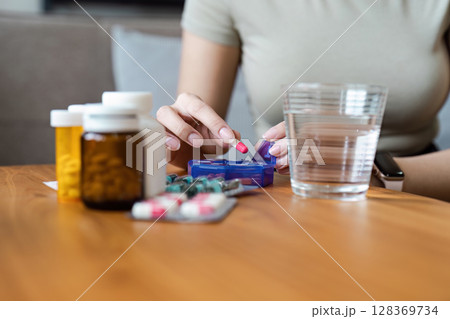 Healthcare at Home: Woman Organizing Pills with Medication Bottles and Water Glass on Wooden Table for Daily Health Management 128369734