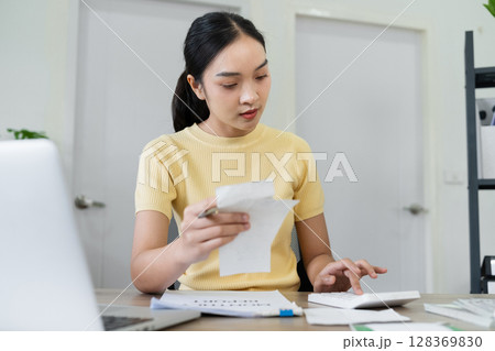 Young Woman Calculating Monthly Bills at Home Using a Calculator and Reviewing Receipts 128369830