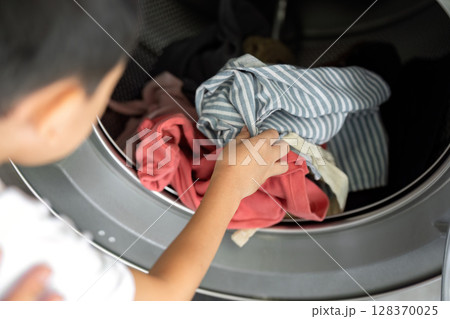 A young boy carefully places clothes into a washing machine, learning about laundry and the importance of cleanliness.. 128370025