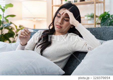 Young Woman Checking Thermometer While Resting on Couch, Experiencing Fever and Illness at Home Young Woman Checking Thermometer While Resting on Couch, Experiencing Fever and Illness at Home 128370054