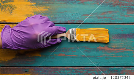 Woman hand in purple rubber glove painting wooden surface with yellow paint brush, creative home renovation and DIY project with tools 128375342