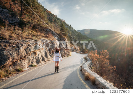 Woman road mountain. A woman in a white sweater, black boots and a hat walks along a winding alpine path between the mountains at sunset in late summer. The concept of travel. 128377606