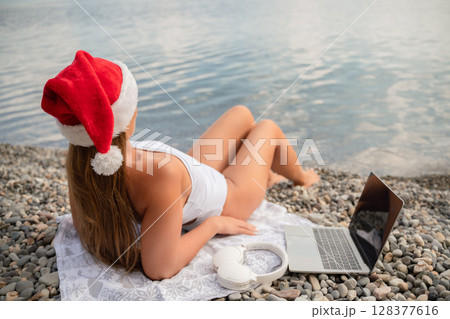 A woman is laying on the beach with a laptop and headphones on her head. She is wearing a red Santa hat. 128377616