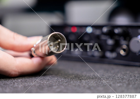 Human hand holds an XLR audio cable connector, poised for connection to a blurred professional sound interface, showcasing audio engineering and recording 128377887