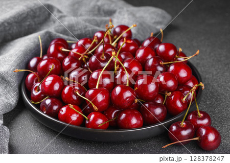 White plate full of fresh ripe red sweet cherry and linen napkin on black texured bakground 128378247