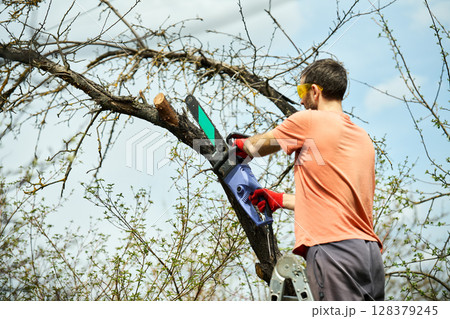 Young man cutting trees using electrical chainsaw in motion at his backyard 128379245