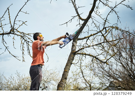 Young man cutting trees using electrical chainsaw in motion at his backyard Young man cutting trees using electrical chainsaw in motion at his backyard 128379249