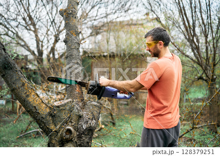 Young man cutting trees using electrical chainsaw in motion at his backyard Young man cutting trees using electrical chainsaw in motion at his backyard 128379251