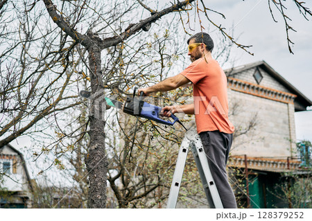 Young man cutting trees using electrical chainsaw in motion at his backyard 128379252