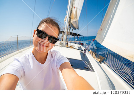 Happy young woman sitting on the bow of a sailing yacht with full sails near Croatia. Freedom, adventure, summer lifestyle and slow travel at sea. 128379335