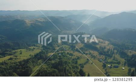 Aerial view of a fog creeping over the mountains surrounding the valley on a summer day in the morning. Aerial drone view of houses in a mountain valley on a summer day. 128381295