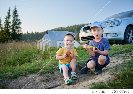 Two young boys sit on grassy hillside, enjoying snacks. Boy in yellow shirt holds bun, while boy in blue shirt and rain boots eats corn on cob. In background tall pine trees under clear blue sky. Two young boys sit on grassy hillside, enjoying snacks. Boy in yellow shirt holds bun, while boy in blue shirt and rain boots eats corn on cob. In background tall pine trees under clear blue sky. 128385567