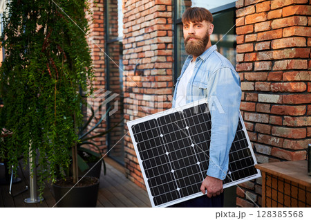 Portrait of bearded man holds solar panel, standing in front of brick building and lush greenery. Guy in shirt and t-shirt exudes calm and eco-conscious vibe, blending nature with urban elements. Portrait of bearded man holds solar panel, standing in front of brick building and lush greenery. Guy in shirt and t-shirt exudes calm and eco-conscious vibe, blending nature with urban elements. 128385568