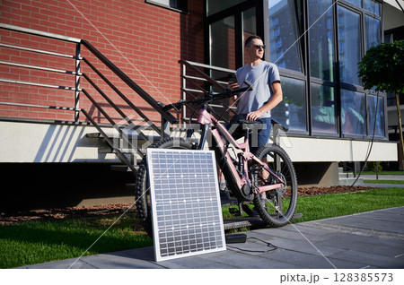 Pink electric mountain bike connected to charging device with solar panel. Man cyclist stands on staircase beside brick building, using smartphone. 128385573
