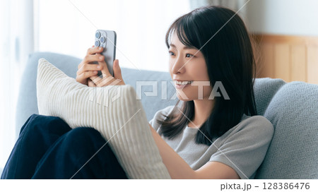 ソファに座ってスマートフォンを使う笑顔の女性 ソファに座ってスマートフォンを使う笑顔の女性 128386476