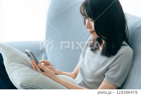 ソファに座ってスマートフォンを使う笑顔の女性 ソファに座ってスマートフォンを使う笑顔の女性 128386478