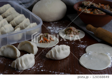 Several raw pork gyoza dumplings being folded on floured wooden table dough circles and filling in progress visible during Japanese cooking at home traditional meal preparation view Several raw pork gyoza dumplings being folded on floured wooden table dough circles and filling in progress visible during Japanese cooking at home traditional meal preparation view 128388649