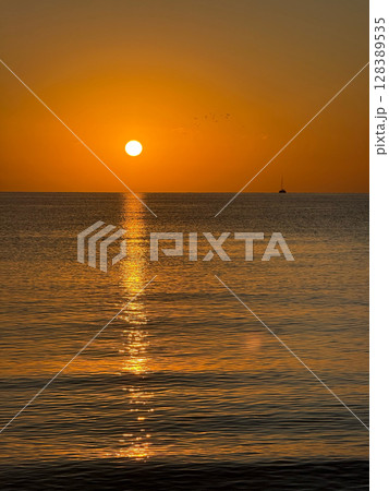 A breathtaking sunset over the calm sea with a sailboat on the horizon. Warm orange and golden tones fill the sky and reflect on the water. Travel, nature, relaxation, and maritime concept A breathtaking sunset over the calm sea with a sailboat on the horizon. Warm orange and golden tones fill the sky and reflect on the water. Travel, nature, relaxation, and maritime concept 128389535