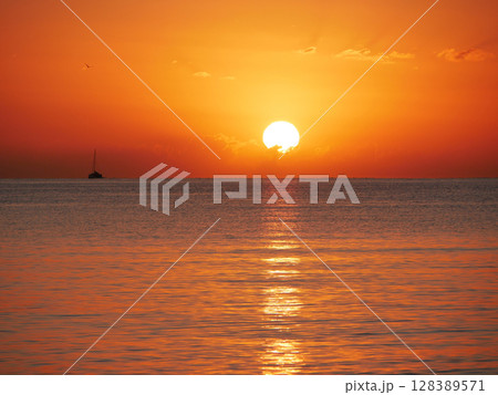 A breathtaking sunset over the calm sea with a sailboat on the horizon. Warm orange and golden tones fill the sky and reflect on the water. Travel, nature, relaxation, and maritime concept 128389571