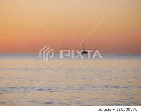 A breathtaking sunset over the calm sea with a sailboat on the horizon. Warm orange and golden tones fill the sky and reflect on the water. Travel, nature, relaxation, and maritime concept 128389576