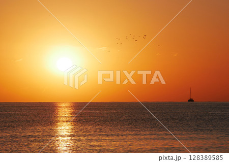 A breathtaking sunset over the calm sea with a sailboat on the horizon. Warm orange and golden tones fill the sky and reflect on the water. Travel, nature, relaxation, and maritime concept A breathtaking sunset over the calm sea with a sailboat on the horizon. Warm orange and golden tones fill the sky and reflect on the water. Travel, nature, relaxation, and maritime concept 128389585
