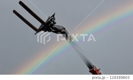 Forks of an automatic forklift reach towards a colorful rainbow in the sky, contrasting industrial machinery with nature's beauty against a gray backdrop. 128389603