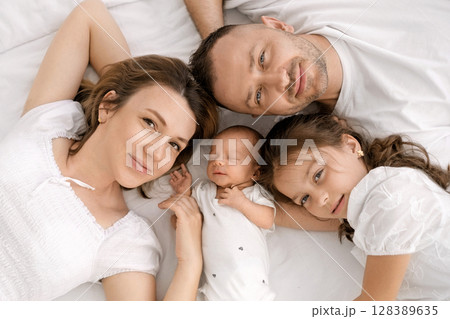 Father, mother, newborn baby and his little sister are lying on the bed looking at the camera, top view Father, mother, newborn baby and his little sister are lying on the bed looking at the camera, top view 128389635