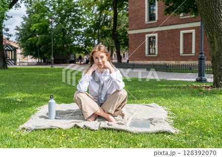 Happy woman lounging barefoot on throw blanket in grassy park, hydrating and smiling to camera  128392070