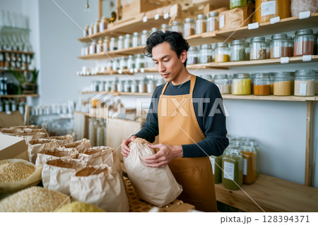 A man in a yellow apron is in a store with many jars and bags of food 128394371