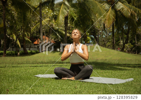 A woman meditates peacefully on a yoga mat under palm trees 128395829