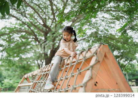 Young Girl Climbing Rope Structure at Playground Under Shady Trees, Engaging in Outdoor Play and Adventure, Joyful Childhood Moments, Natural Environment Young Girl Climbing Rope Structure at Playground Under Shady Trees, Engaging in Outdoor Play and Adventure, Joyful Childhood Moments, Natural Environment 128399014