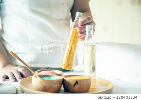 Close-up of hand holding amber bottle of essential oil over wooden tray with colorful spa salts and jars; preparation for beauty wellness massage and holistic skincare treatment Close-up of hand holding amber bottle of essential oil over wooden tray with colorful spa salts and jars; preparation for beauty wellness massage and holistic skincare treatment 128401234
