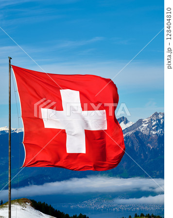Switzerland flag waving against mountain backdrop in bright blue sky with clouds during daytime Switzerland flag waving against mountain backdrop in bright blue sky with clouds during daytime 128404810